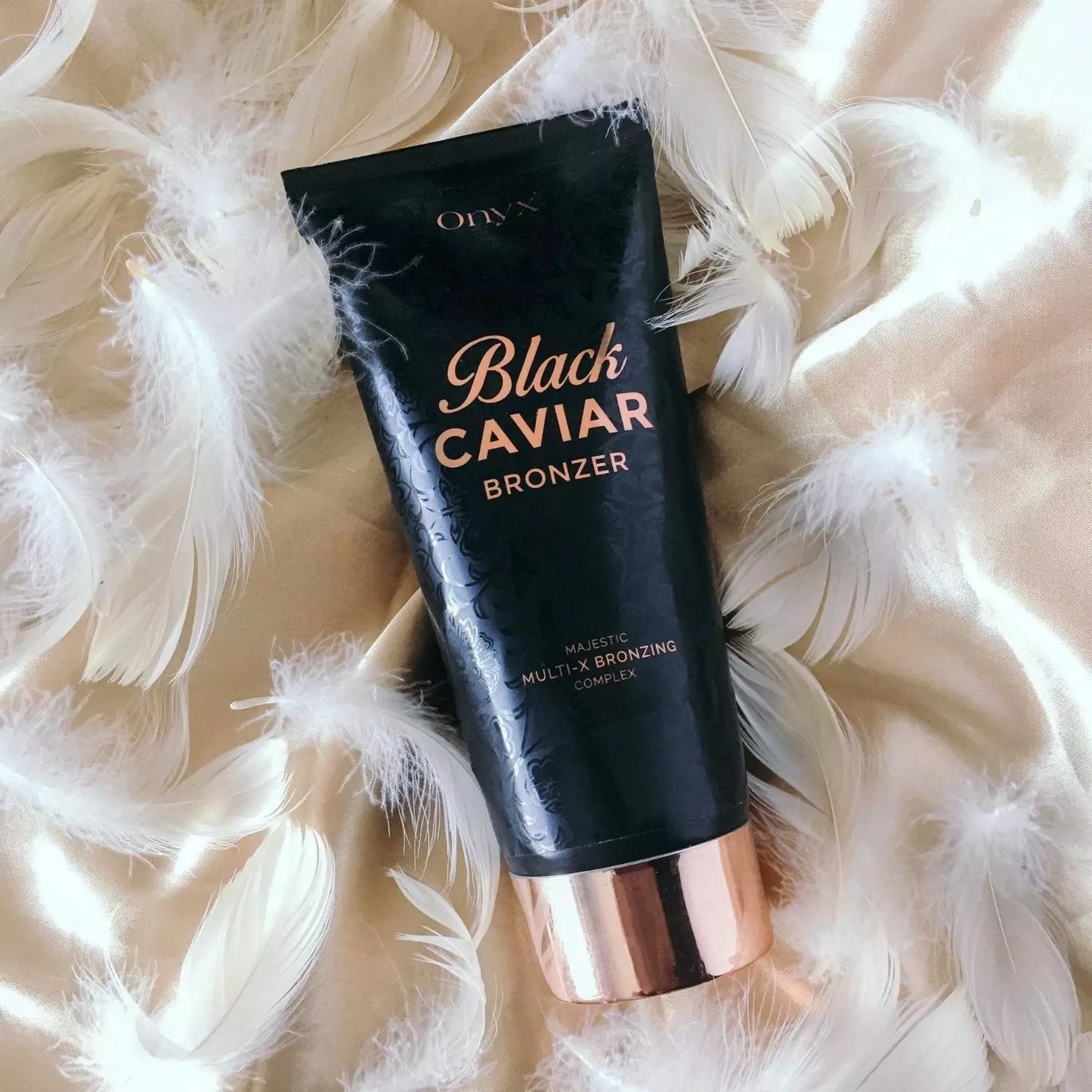Onyx Black Caviar Bronzer położony na złotej satynie z białymi piórami; elegancki bronzer do opalania zapewniający luksusowy efekt skóry muśniętej słońcem