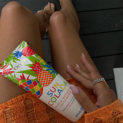 Kobieta w pomarańczowym swetrze trzyma balsam Onyx Suncolada na udach, widoczna gładka i opalona skóra nóg oraz pierścionek z kryształkami