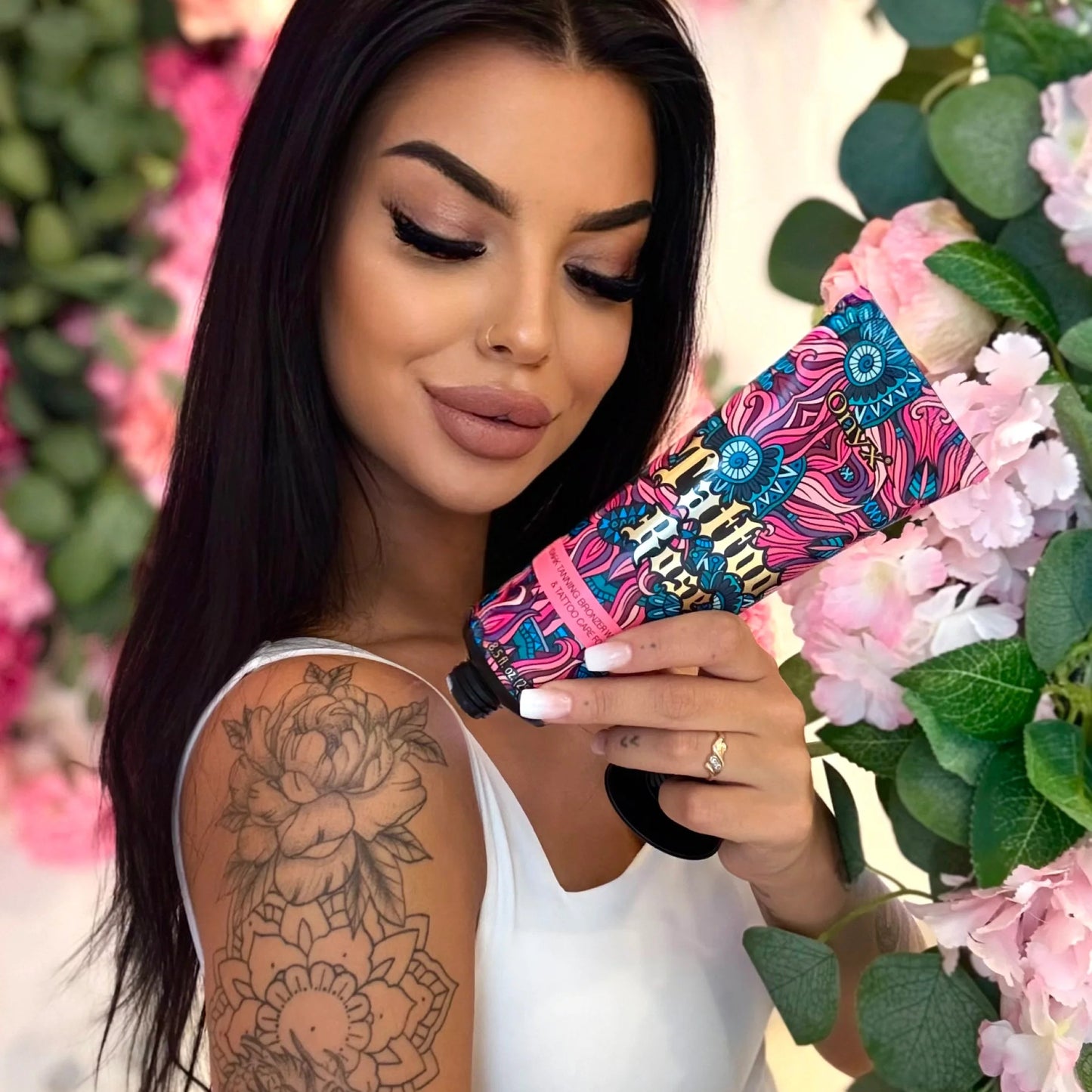 Kobieta w białym topie trzyma tubę Onyx Tattoo Rose na tle różowych kwiatów i zielonych liści.