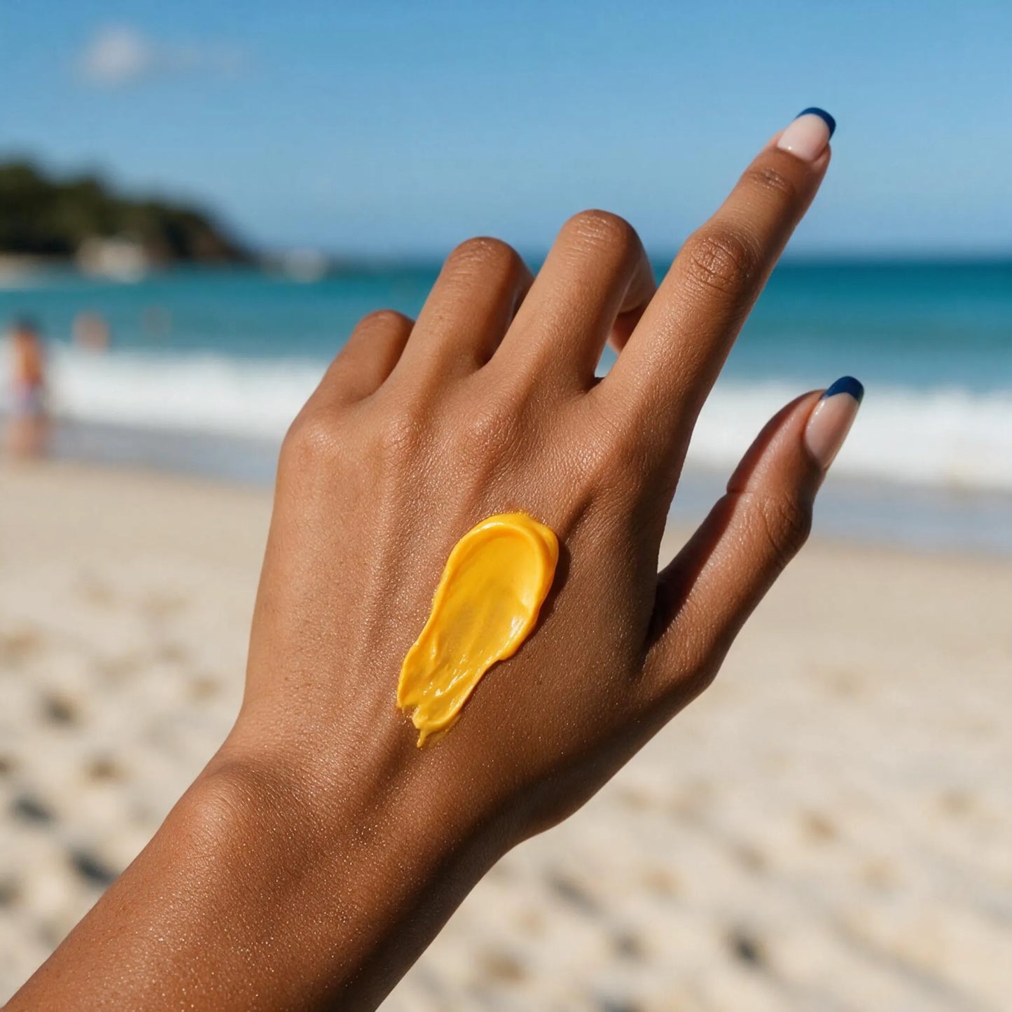 A close-up of a person's hand with a dollop of bright yellow sunscreen cream on the back, set against a blurred beach background with blue ocean and sky.